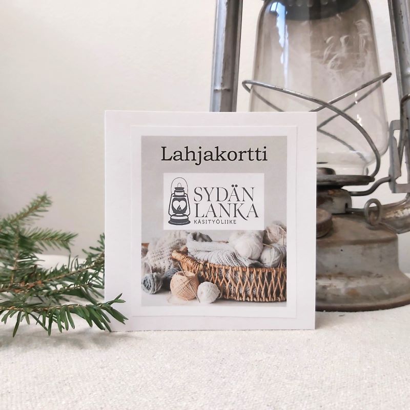 Käsityöliike Sydänlangan lahjakortti ja taustalla havuja ja harmaa myrskylyhty